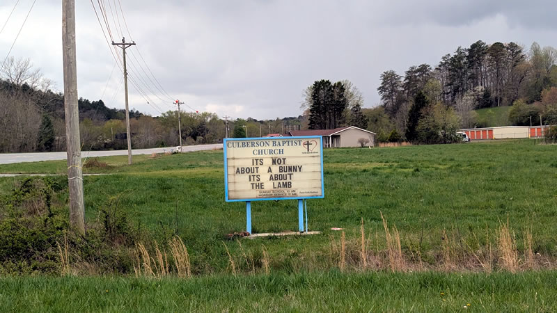 church sign in a grass field with self storage units in the background