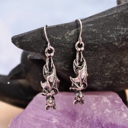 Pair of silver-tone three-dimensional bat drop earrings displayed against a dark stone background with a purple crystal.