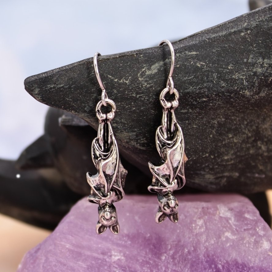 Pair of silver-tone three-dimensional bat drop earrings displayed against a dark stone background with a purple crystal.