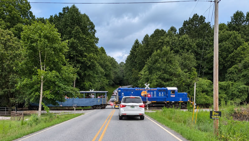 A Blue Ridge Scenic Railway train crosses a rural road surrounded by lush green forests in the Blue Ridge Mountains of Georgia, with a white SUV waiting at the railroad crossing.