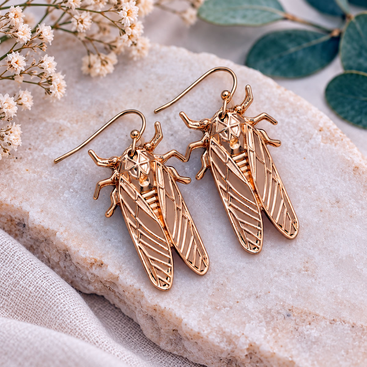 Pair of gold-tone cicada dangle earrings displayed on a natural stone against a soft pink background, showing their detailed wings and elongated insect bodies.