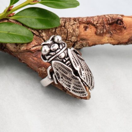 Detailed silver cicada statement ring displayed on rustic wood, showing its sculpted body and intricately patterned wings.