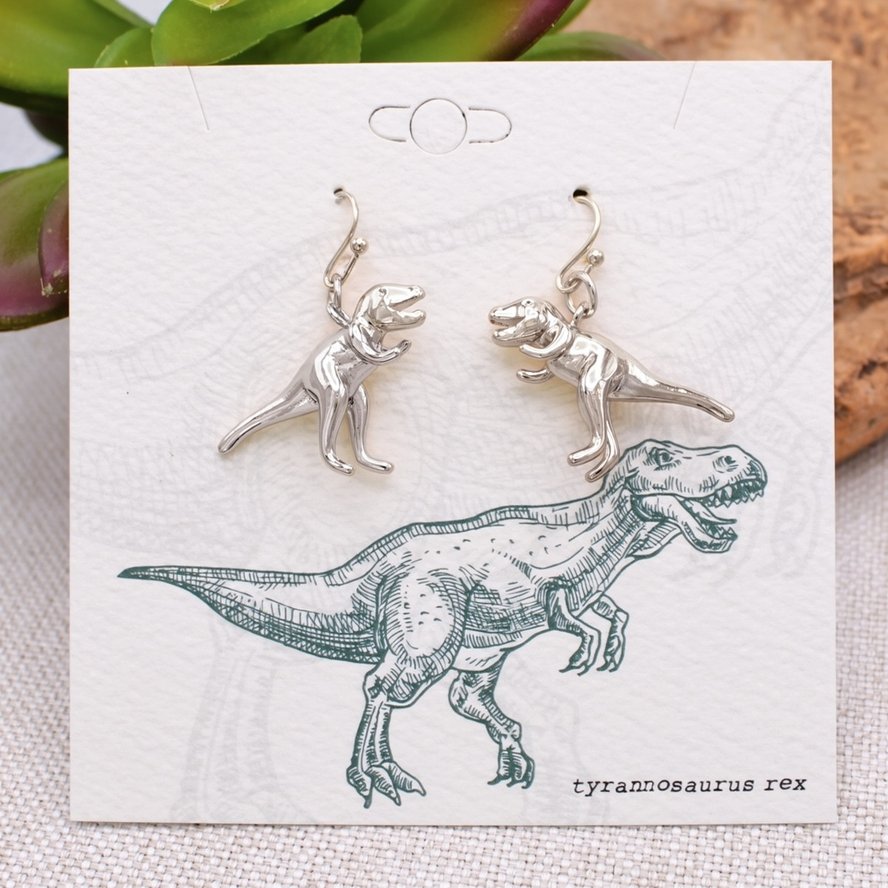 Pair of silver-tone Tyrannosaurus rex dangle earrings displayed on a vintage-style card illustrated with a T. rex dinosaur.