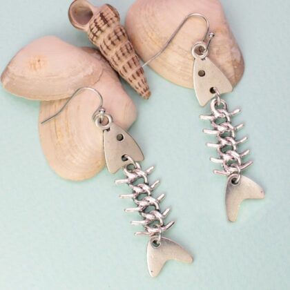 Pair of silver-tone fish bone drop earrings displayed beside seashells on a pale aqua background.