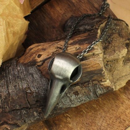 Silver-tone crow skull pendant necklace arranged on rustic wood to showcase its burnished metal finish.
