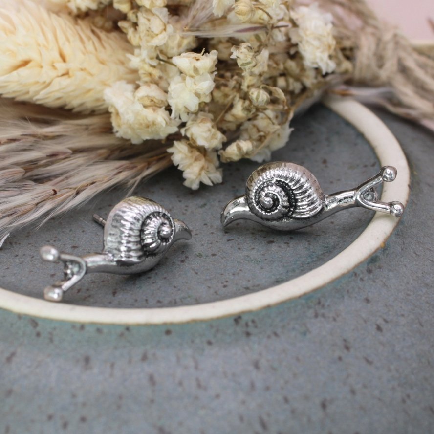 Silver-tone snail earrings displayed on a small ceramic dish with dried flowers, showcasing their whimsical nature-inspired design.