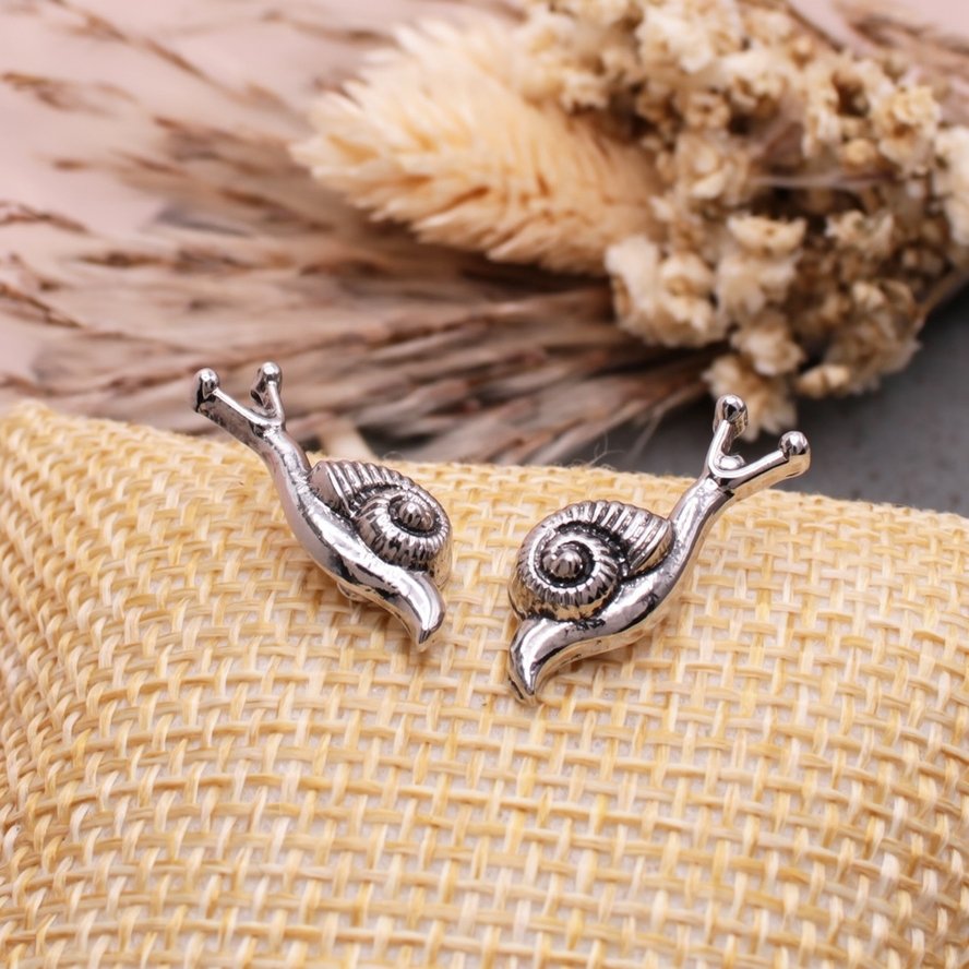 Pair of silver-tone snail stud earrings with detailed spiral shells displayed on a woven natural-texture surface.