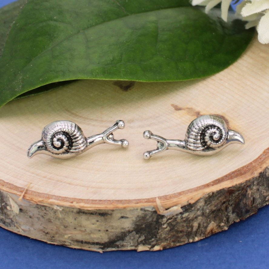 Silver snail post earrings shown side by side on a wood slice, highlighting their spiral shells and tiny antennae.